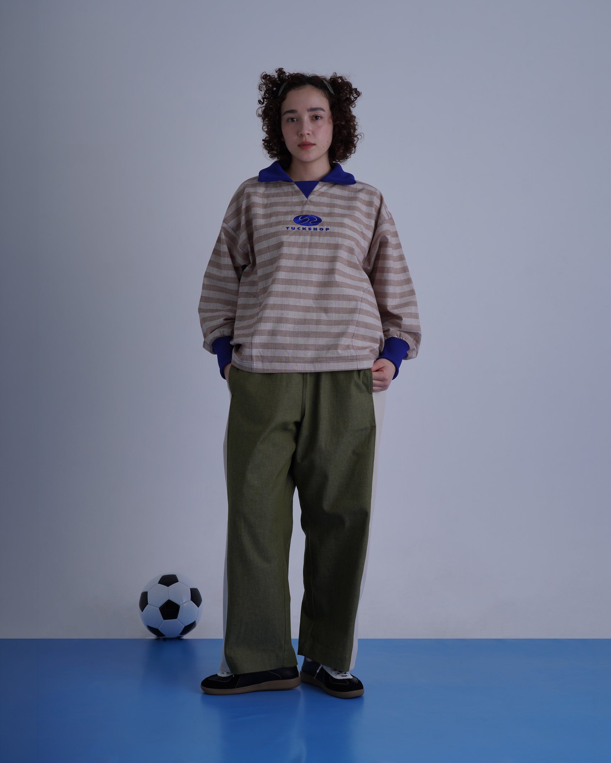 FOOTBALL POLO PULLOVERを着たモデルが、サッカーボールの近くでポーズを取る。ストライプデザインとカラーコントラストが特徴。