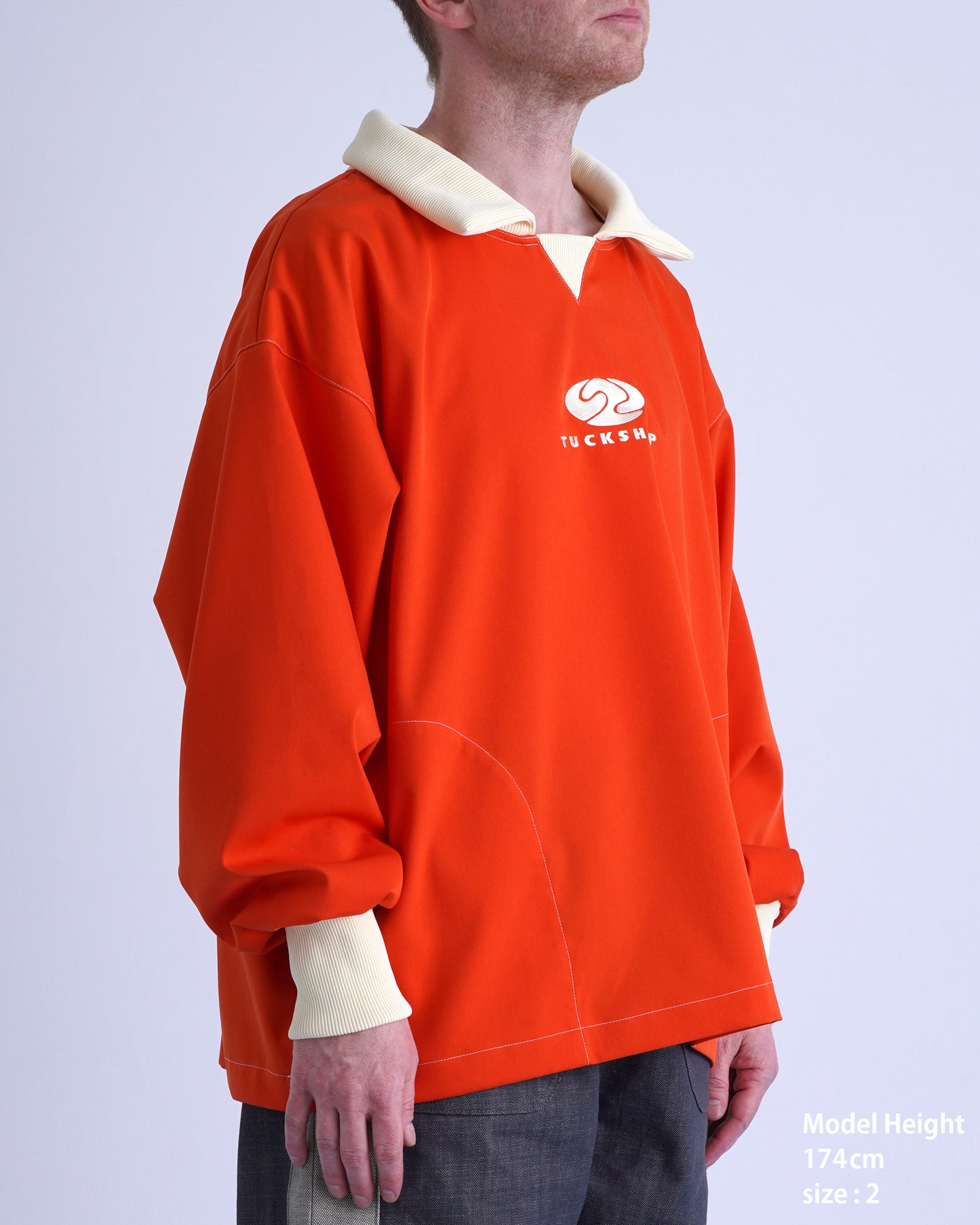 FOOTBALL POLO PULLOVERのオレンジ色プルオーバーを着用したモデル。スプレッドカラーとロゴ刺繍が特徴。