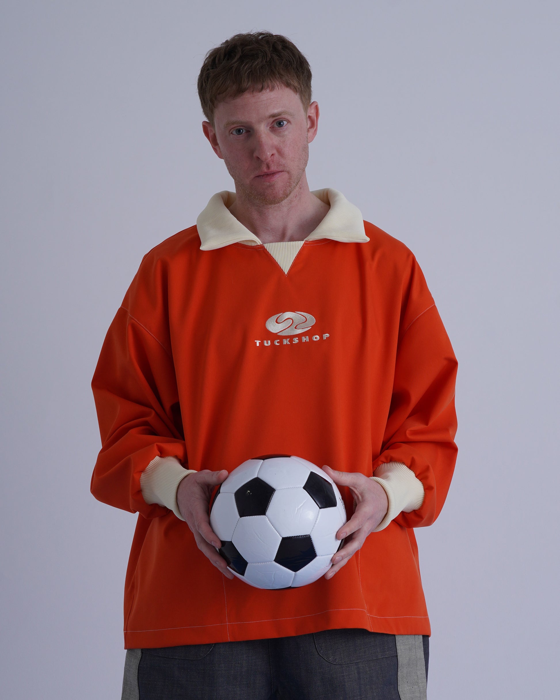 FOOTBALL POLO PULLOVERを着用し、サッカーボールを持つ男性の画像。オレンジ色のプルオーバー。