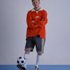 FOOTBALL POLO PULLOVERを着てサッカーボールを持った男性が立っている画像。オレンジ色のプルオーバーと灰色のショートパンツ。