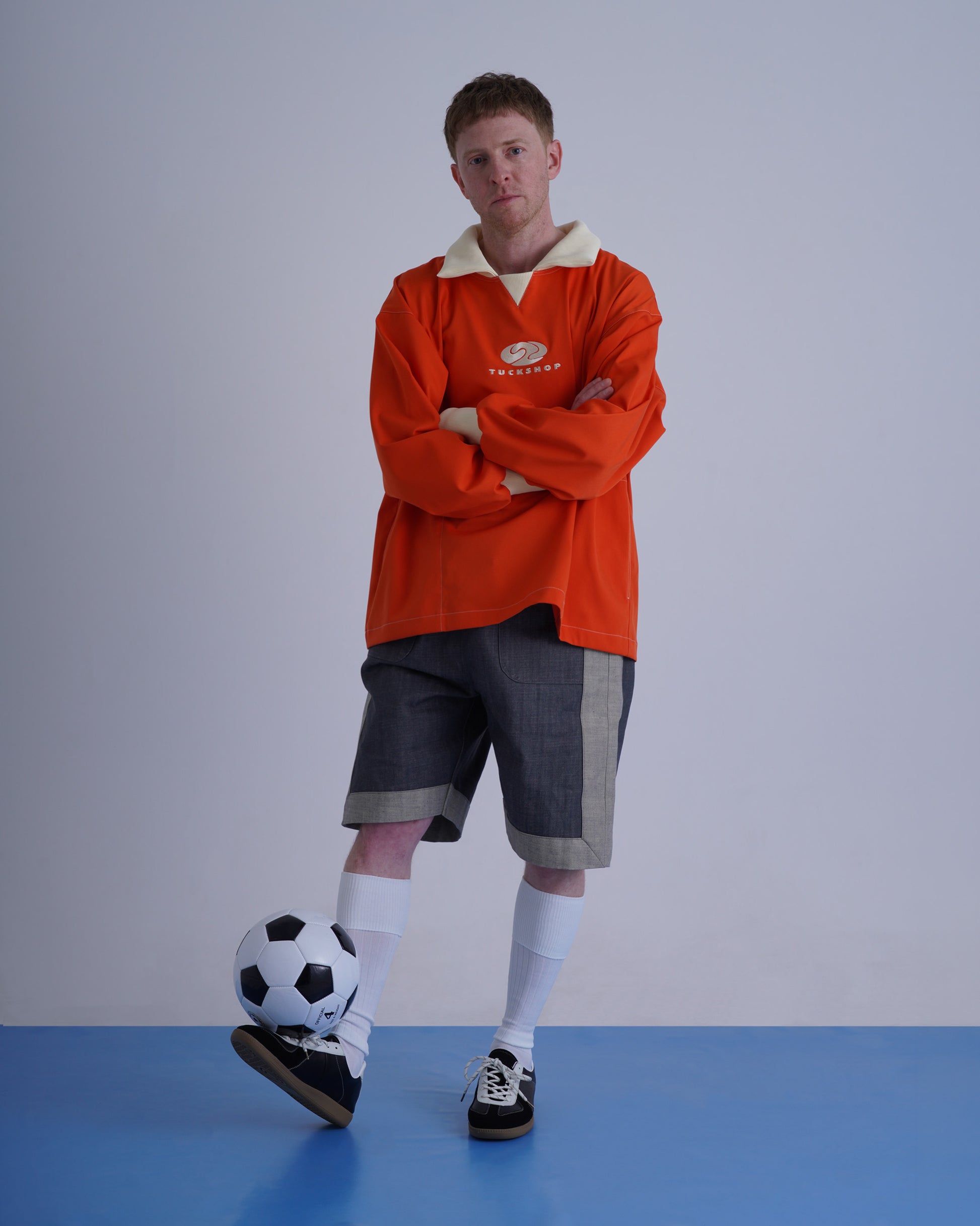 FOOTBALL POLO PULLOVERを着てサッカーボールを持った男性が立っている画像。オレンジ色のプルオーバーと灰色のショートパンツ。