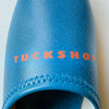 Close-up of blue faux leather slipper with red 'TUCKSHOP' logo print on the instep.