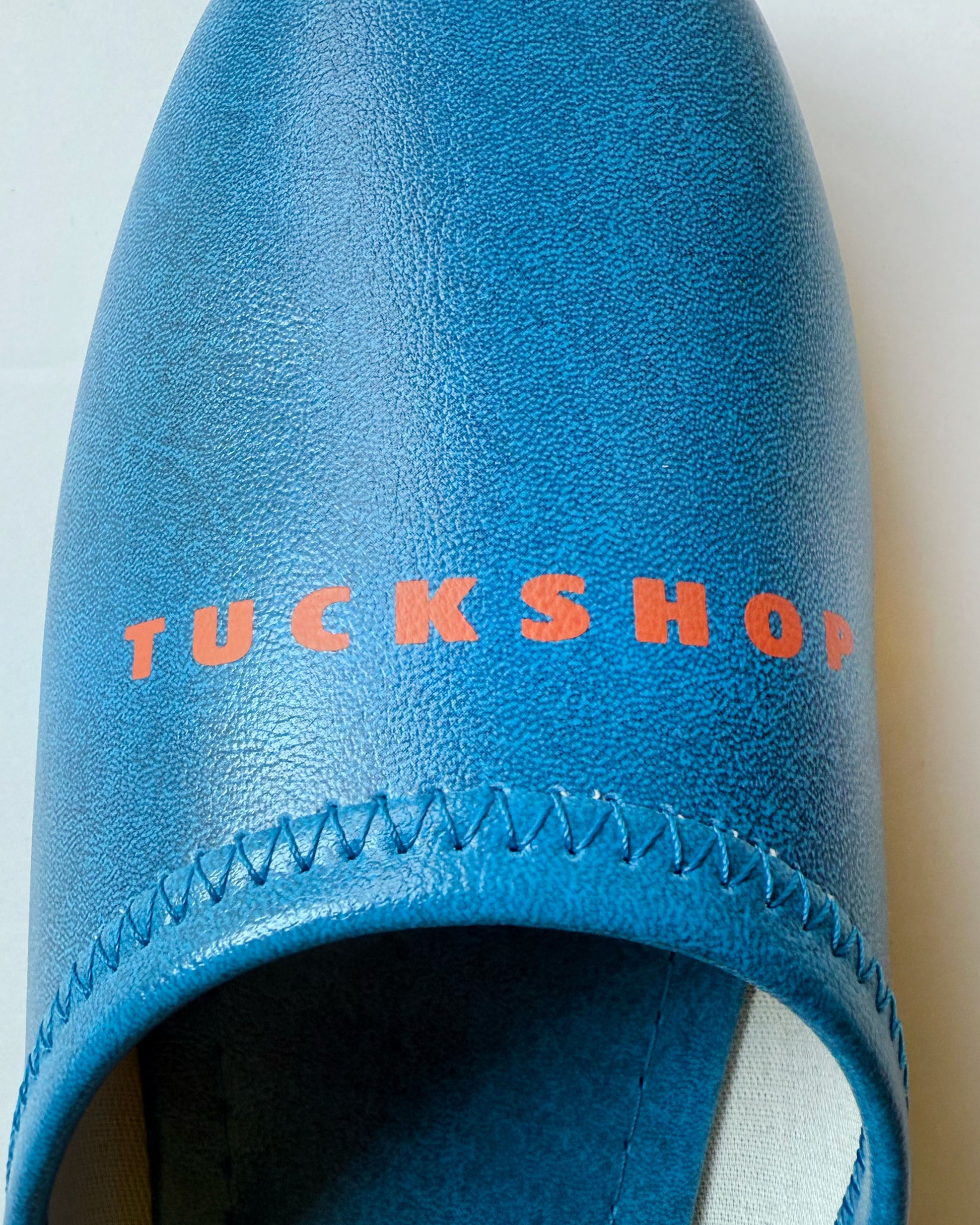 Close-up of blue faux leather slipper with red 'TUCKSHOP' logo print on the instep.