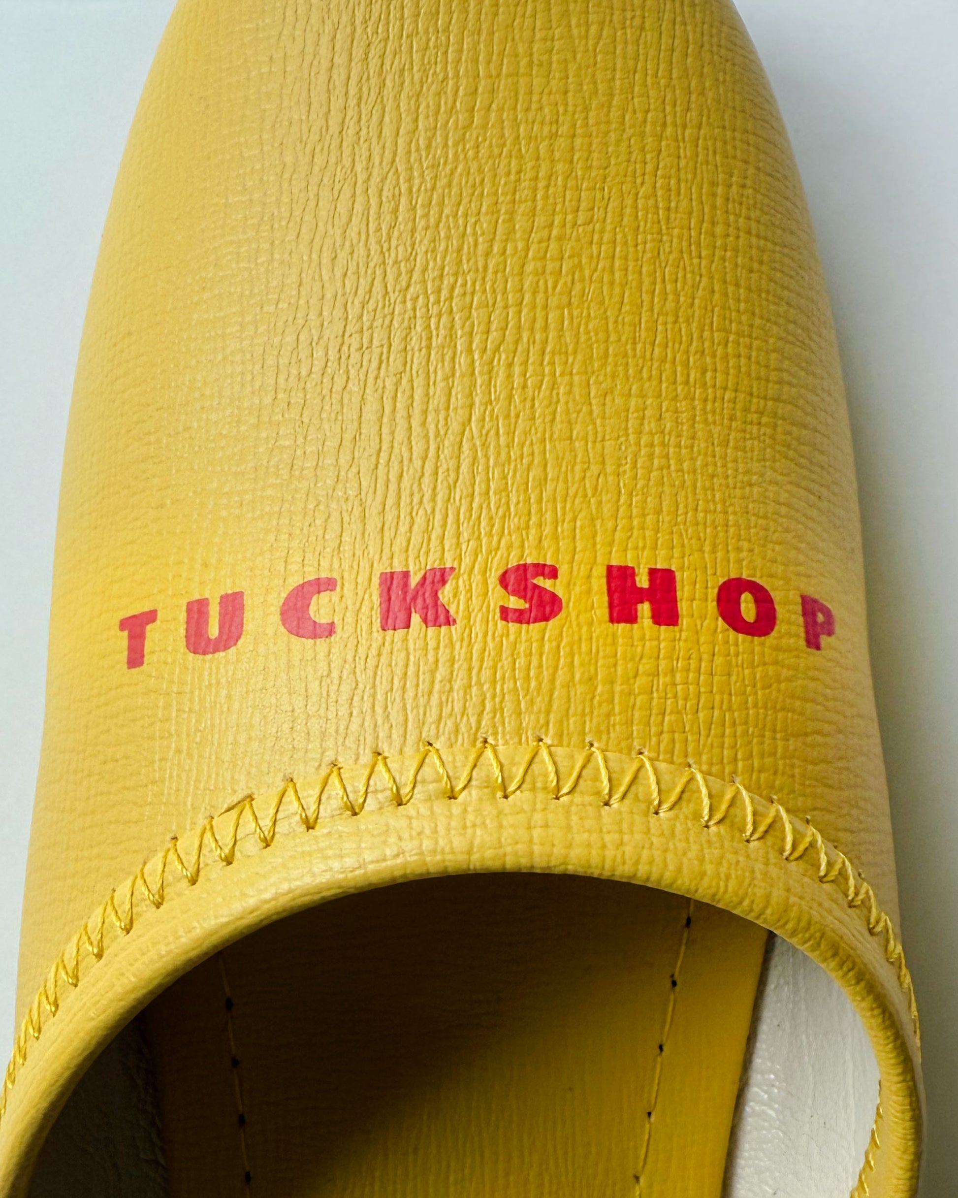 Yellow faux leather slipper with 'TUCKSHOP' logo printed in red on the instep.